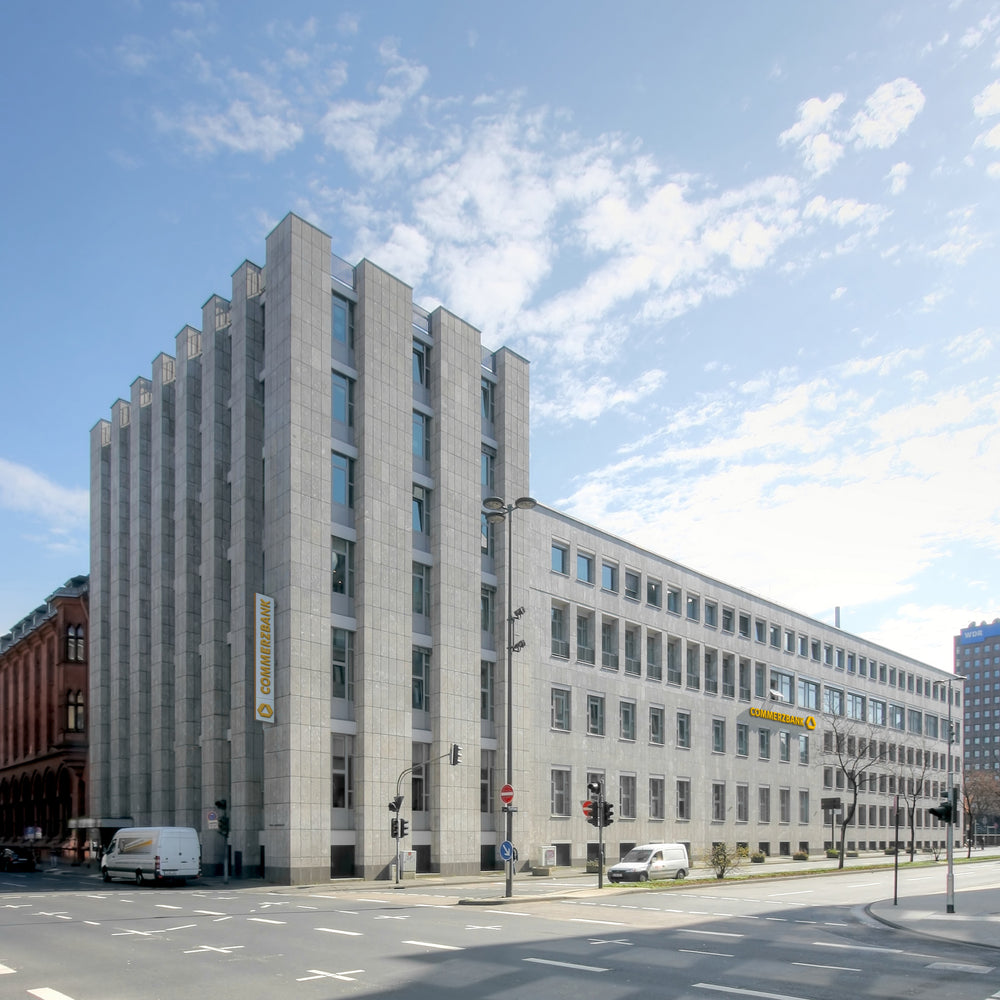 Futuristische Architektur der Commerzbank-Zentrale mit einer Fassade aus geometrischen Natursteinblöcken, die Himmel und städtisches Umfeld reflektieren. Unten auf dem Bild steht 'Welcher Naturstein passt zu Ihnen? Lassen Sie sich inspirieren durch unsere Kunden', was die Wertigkeit von Naturstein in der Unternehmensarchitektur betont.